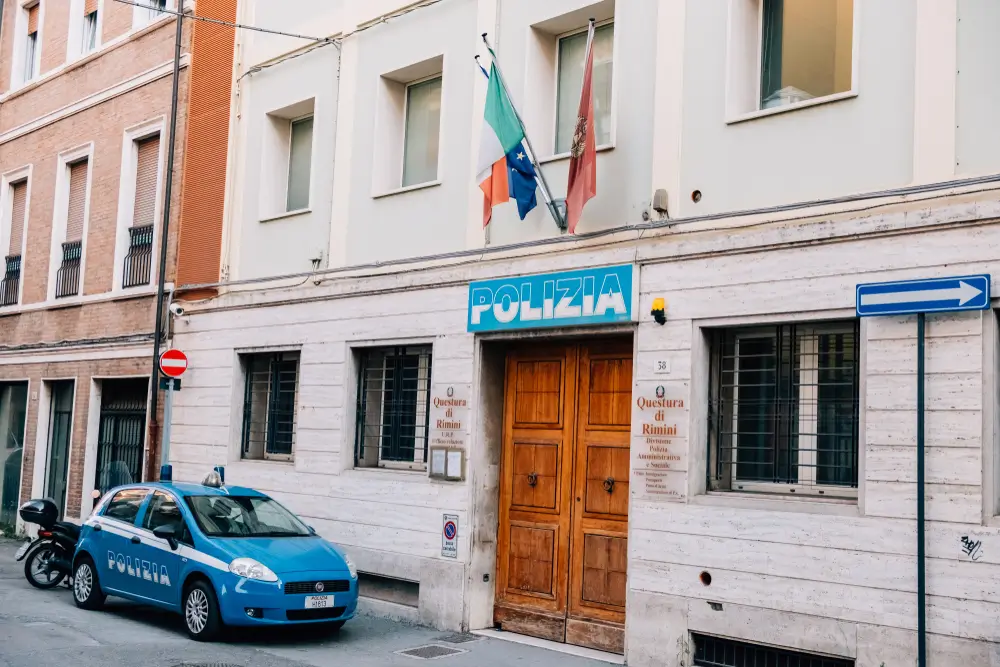Facciata della Questura di Rimini con bandiere istituzionali, insegna Polizia e un'auto della Polizia di Stato.