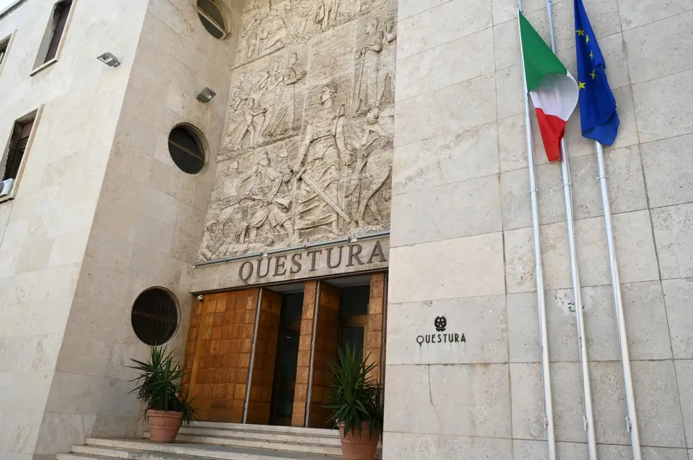 Ingresso della sede di una Questura con bassorilievo in pietra, bandiere istituzionali e insegna della Polizia di Stato.