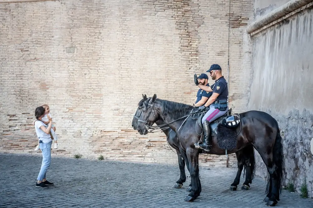 Agenti a cavallo in divisa davanti a un edificio della scuola di polizia salutano una donna con una bambina.