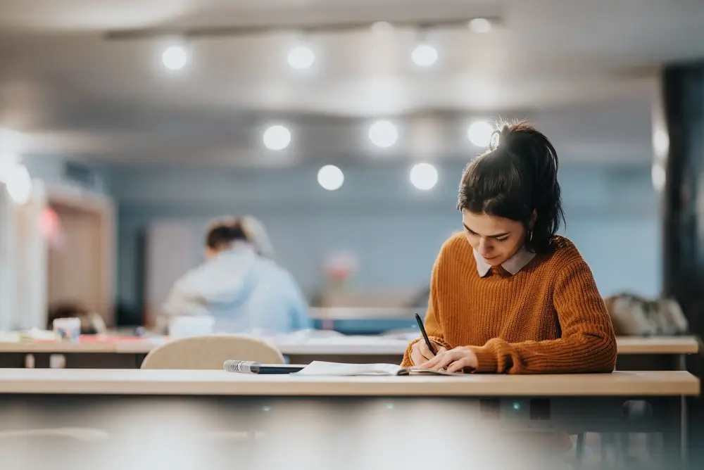 Studentessa che studia per la preparazione del concorso, scrivendo appunti su un quaderno in un'aula studio.